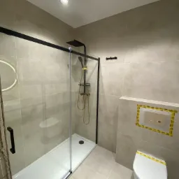 Nowoczesna łazienka z kabiną prysznicową typu walk-in z czarnymi ramami, złotą armaturą i wiszącą toaletą z zabezpieczeniem folią ochronną.