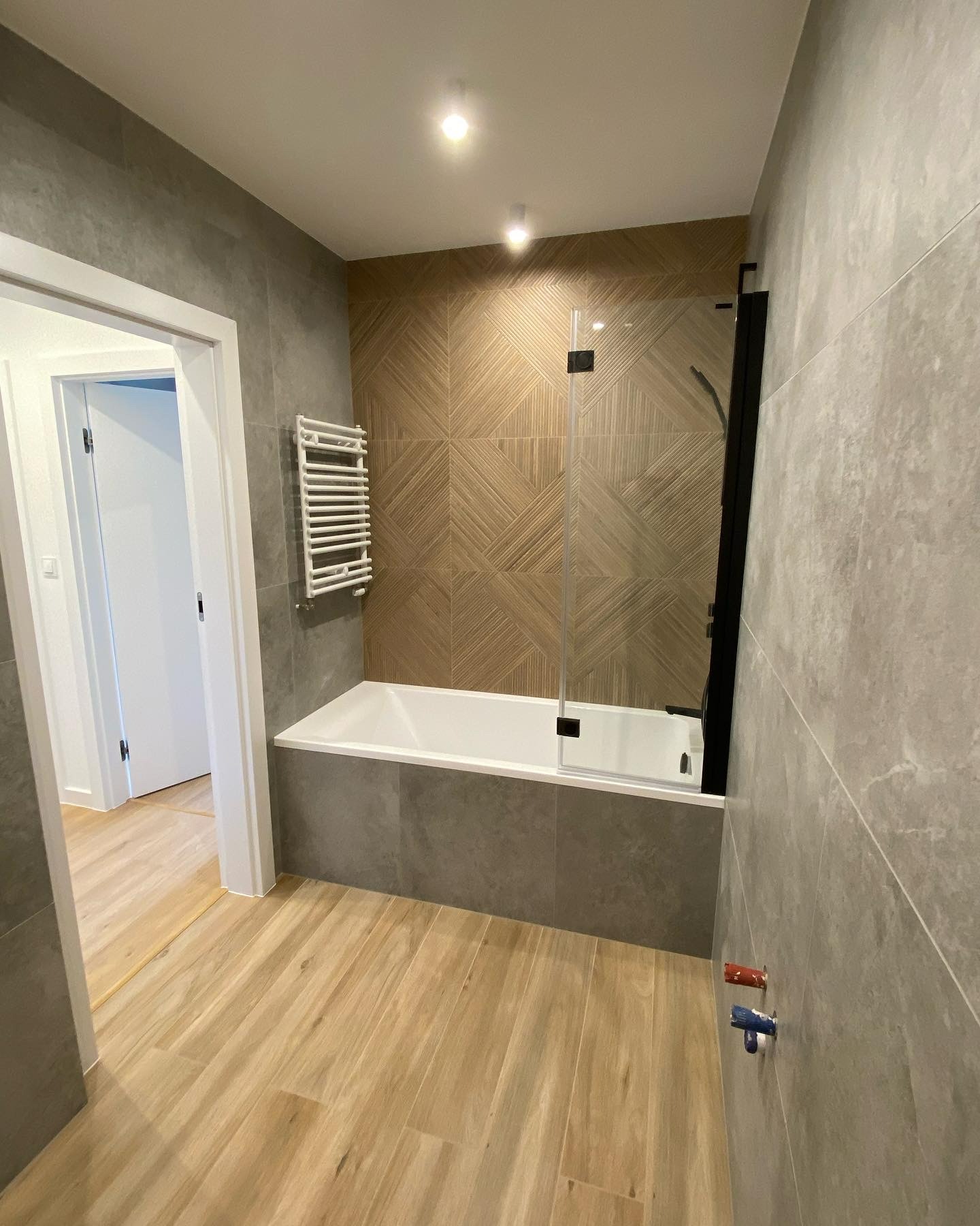 Nowoczesna łazienka po remoncie z płytkami imitującymi drewno na ścianie za wanną i podłodze, szklany parawan nawannowy z czarnymi elementami, grzejnik drabinkowy i szare płytki na pozostałych...