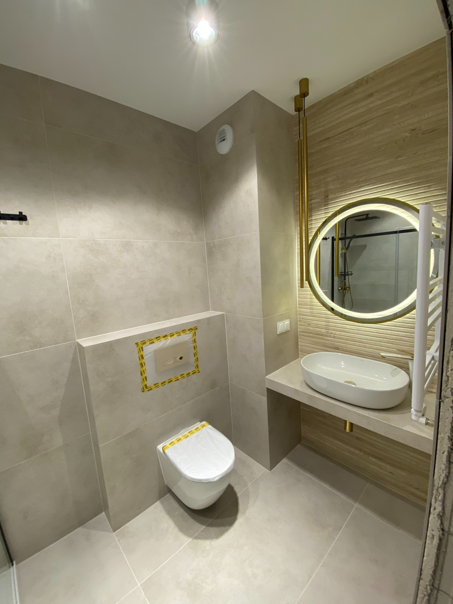 Nowoczesna łazienka w beżowych tonacjach z podświetlanym okrągłym lustrem, wiszącą miską WC z zabezpieczeniem folią, umywalką nablatową na kamiennym blacie i grzejnikiem drabinkowym.