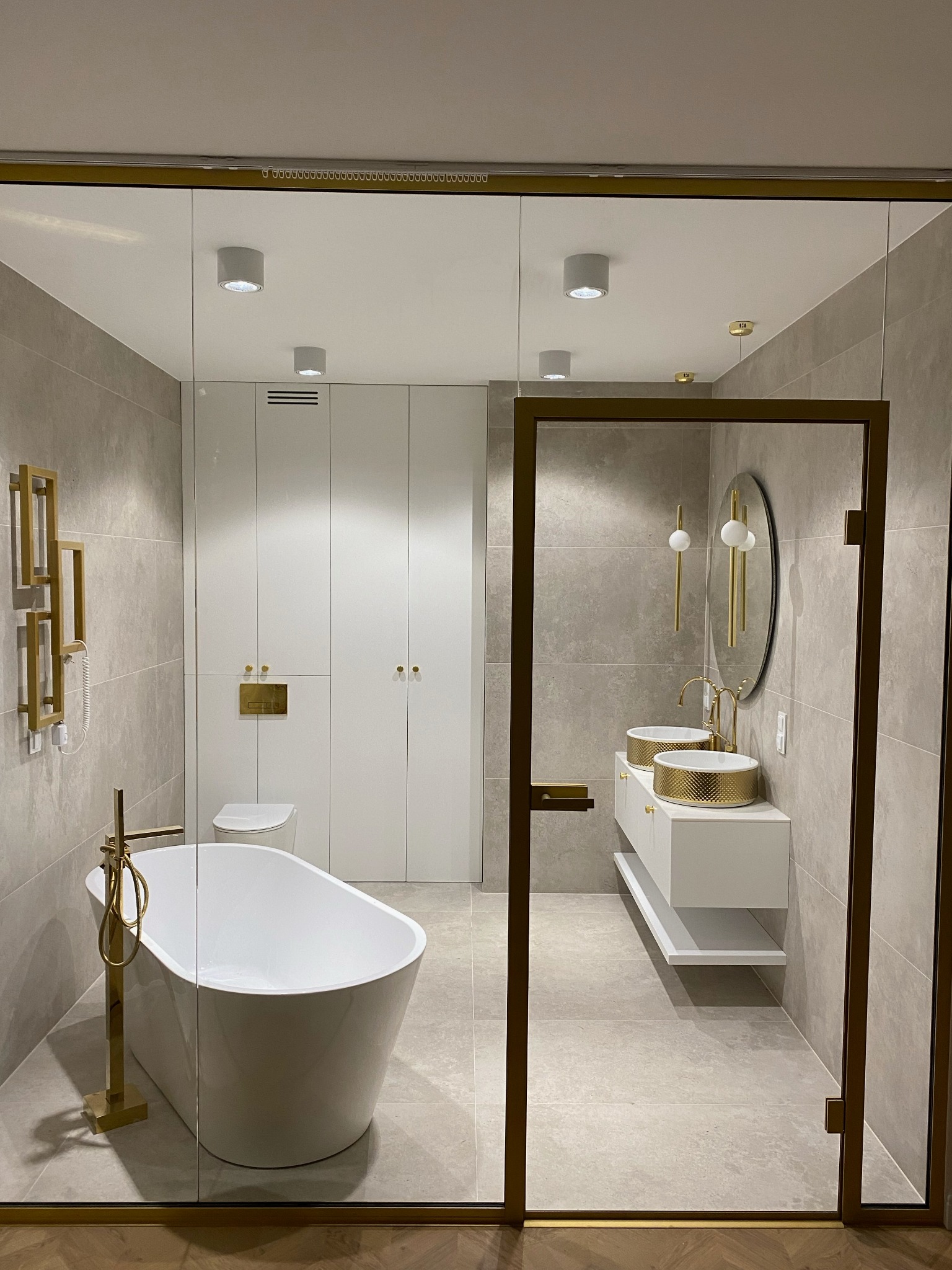 Nowoczesna łazienka z wolnostojącą wanną, podwójną umywalką w złotym kolorze i szklaną kabiną prysznicową z złotymi ramami.