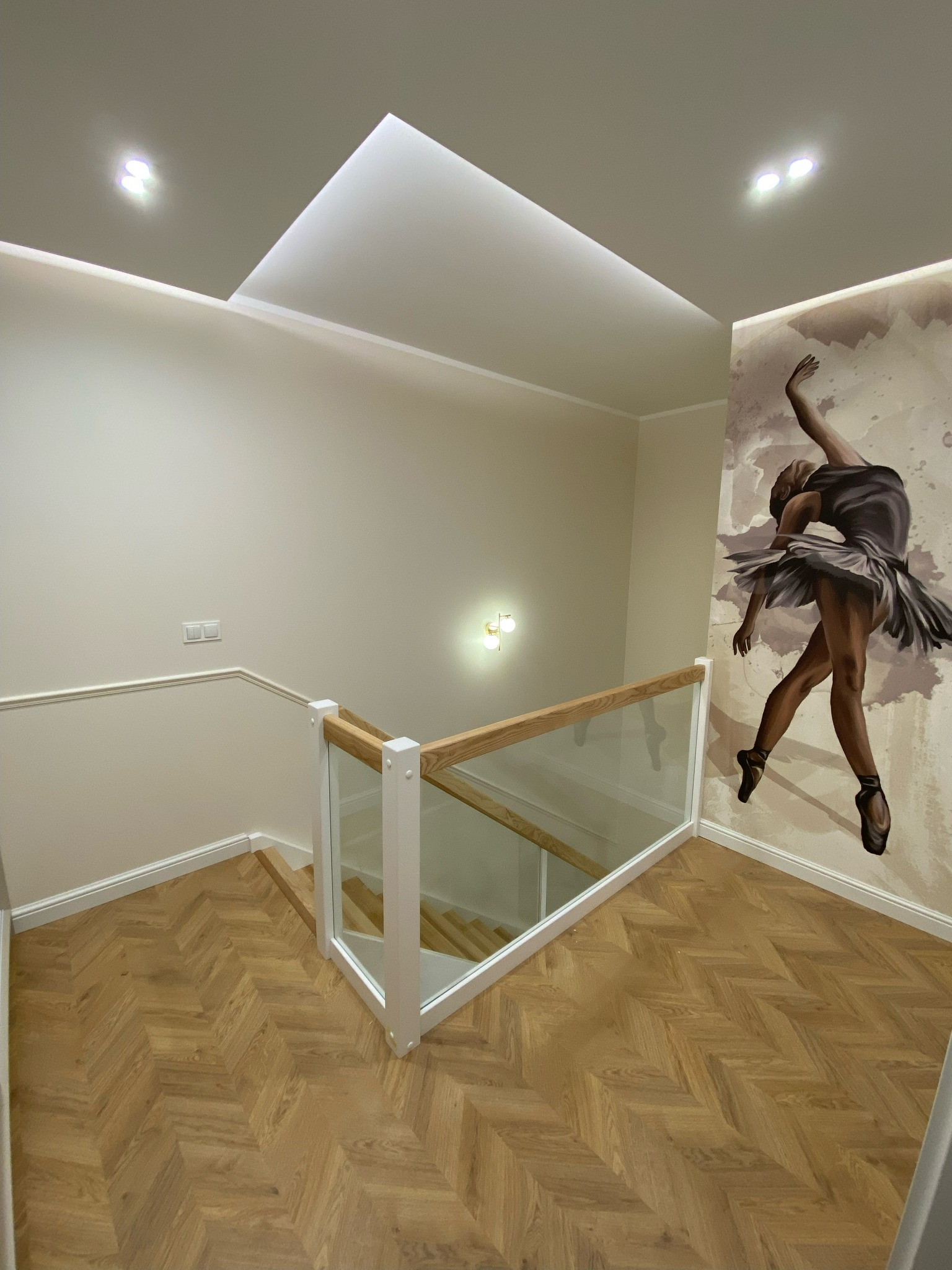Klatka schodowa z parkietem ułożonym w jodełkę, szklana balustrada z drewnianymi elementami, na ścianie mural z baletnicą w sepii, nowoczesne oświetlenie sufitowe.