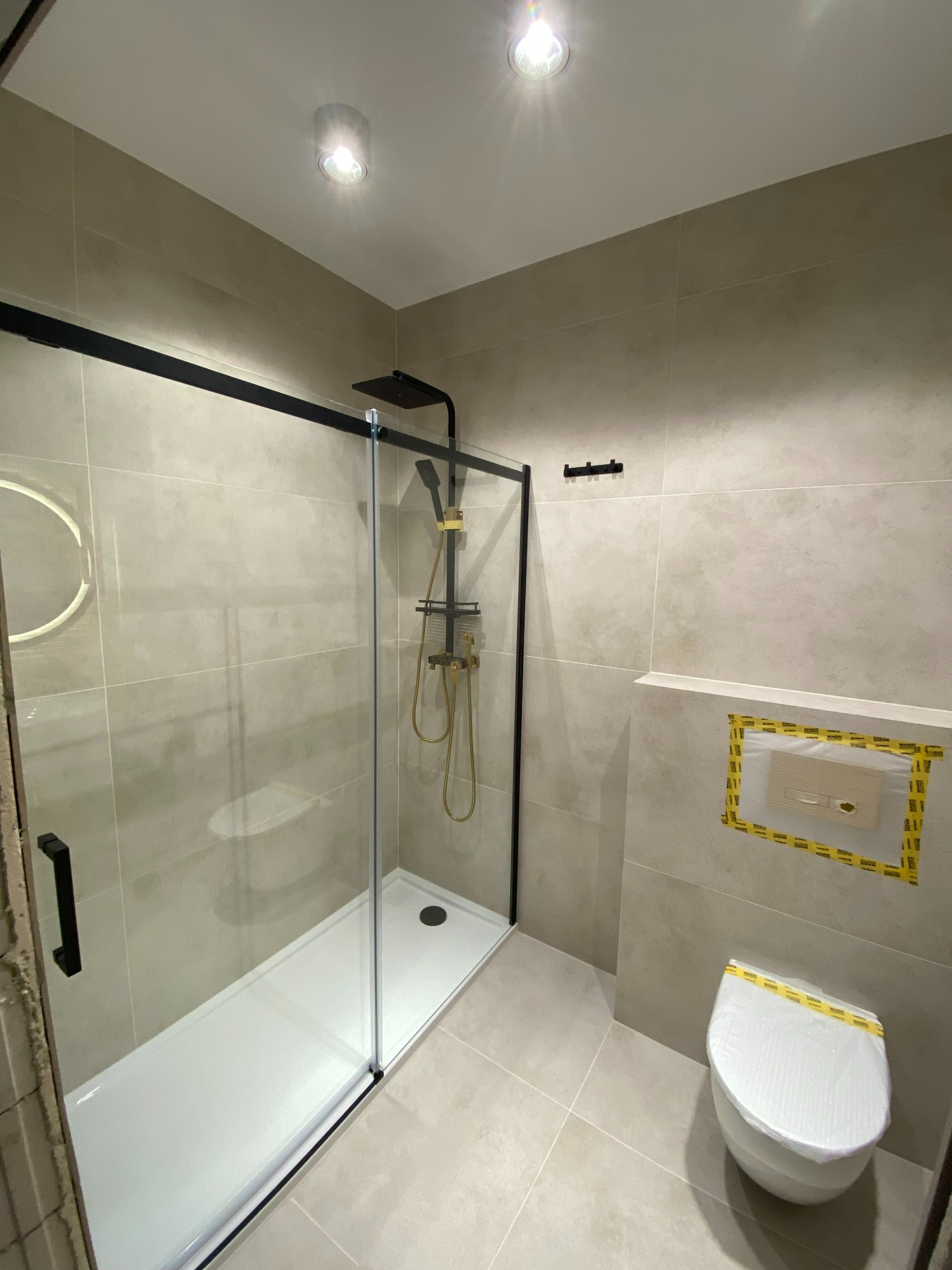 Nowoczesna łazienka z kabiną prysznicową typu walk-in z czarnymi ramami, złotą armaturą i wiszącą toaletą z zabezpieczeniem folią ochronną.