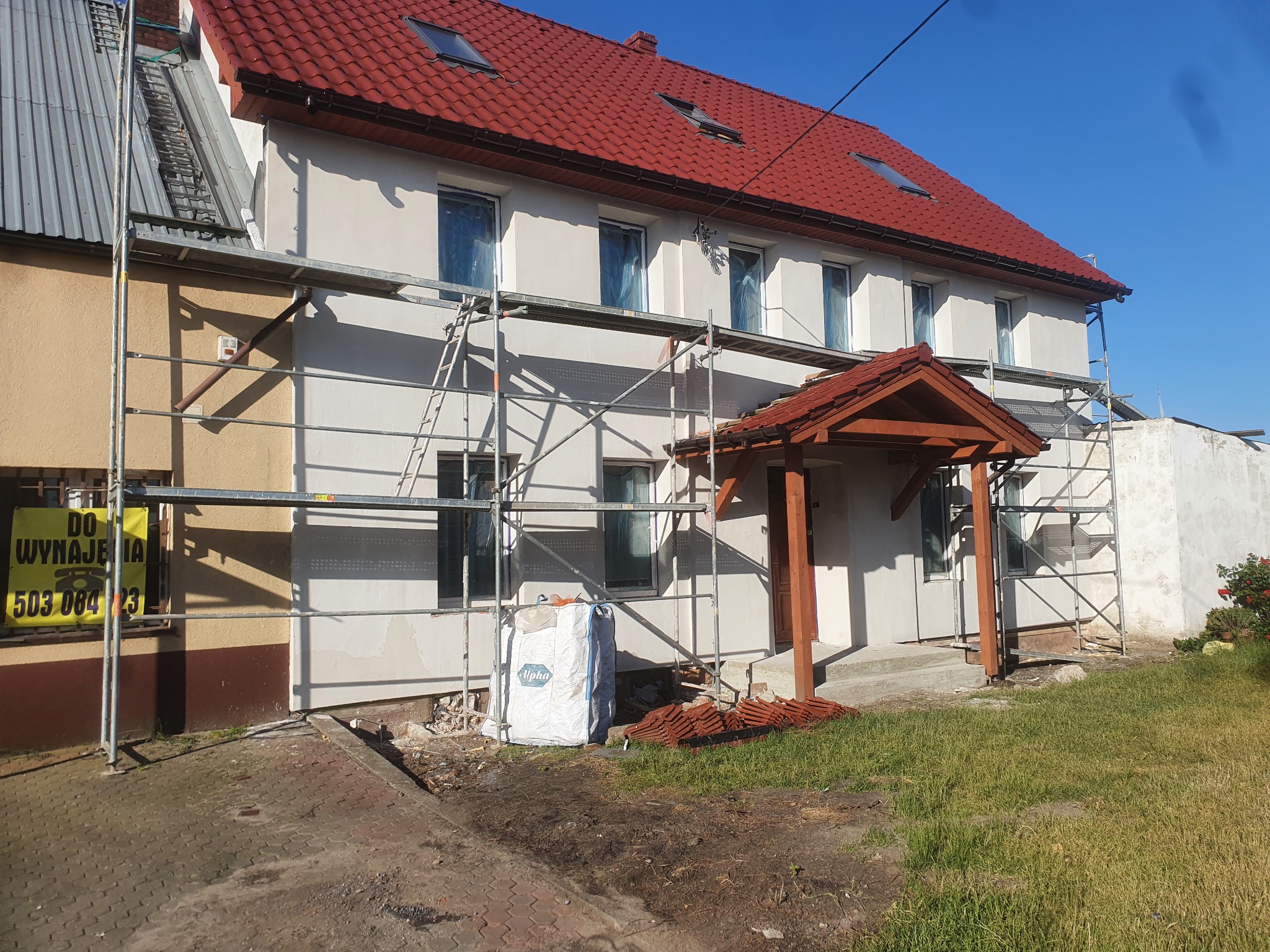 Dom jednorodzinny w trakcie ocieplania elewacji, widoczne rusztowania, zabezpieczone okna folią, biały worek z materiałem izolacyjnym i stos czerwonych dachówek obok wejścia.