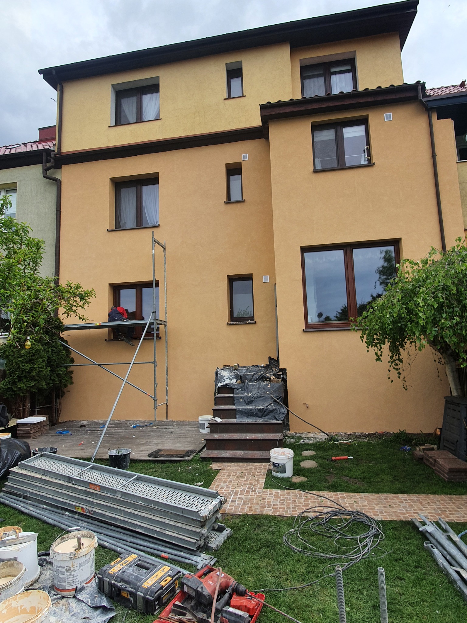 Dom jednorodzinny w Brzesku z elewacją w odcieniach beżu, częściowo osłonięty rusztowaniem, widoczne narzędzia i materiały budowlane na trawniku, pracownik na rusztowaniu przy oknie.