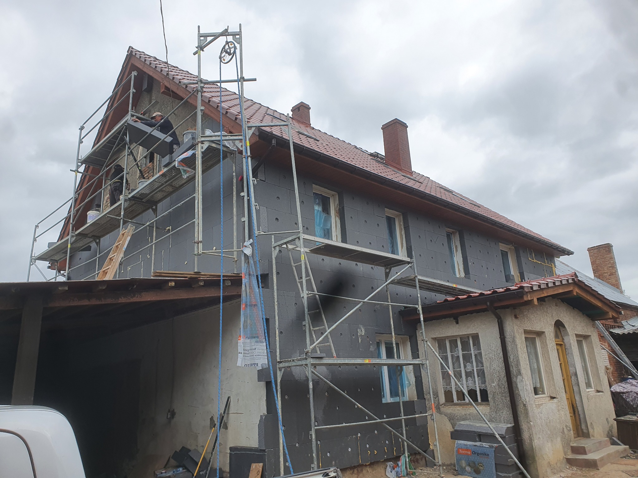 Dwukondygnacyjny dom w trakcie ocieplania grafitowym styropianem, widoczne rusztowania i pracownicy na wysokości, częściowo ocieplona elewacja z widocznymi otworami po kołkach, pochmurne niebo