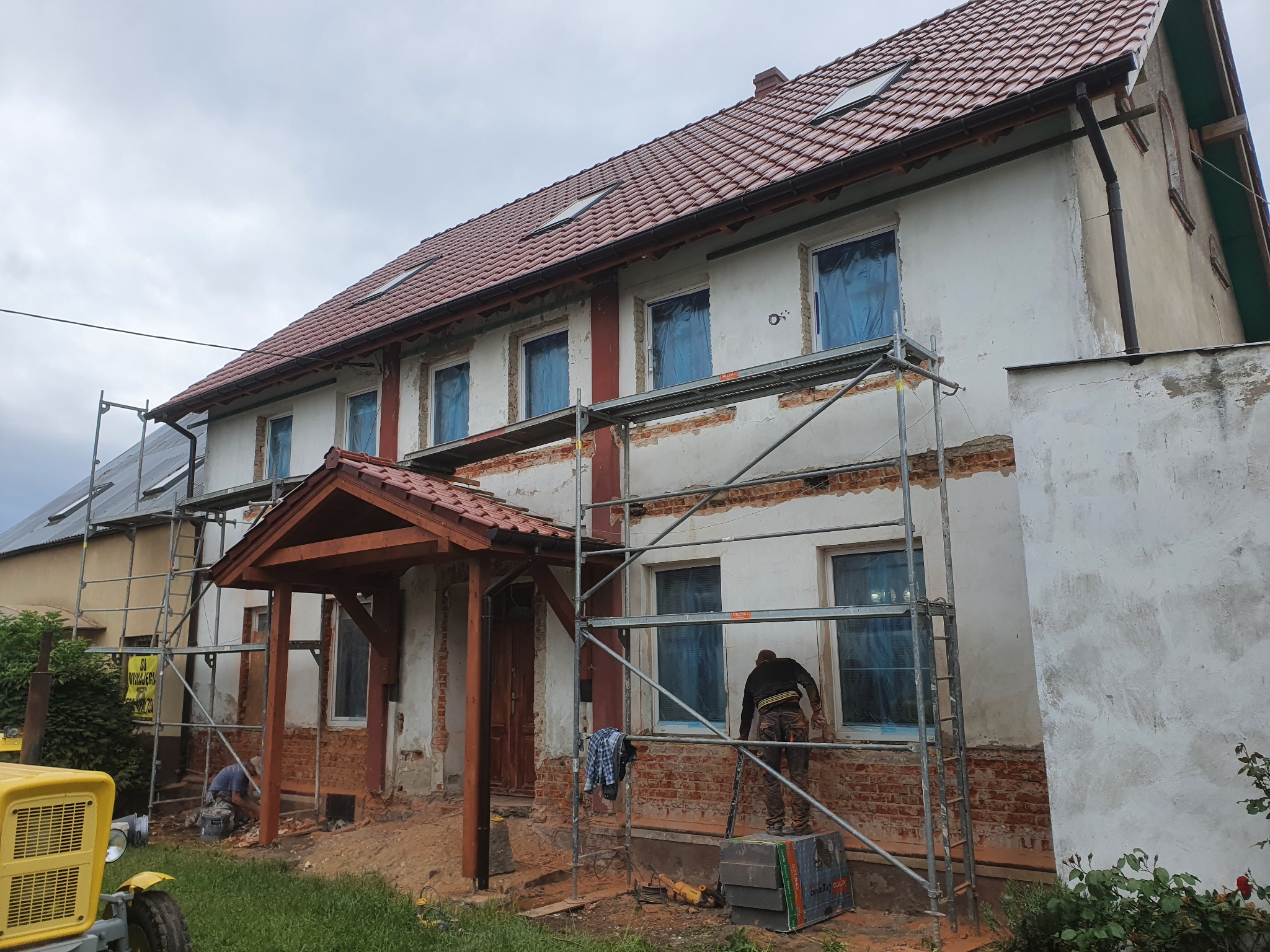 Dwukondygnacyjny dom z czerwonej cegły i białym tynkiem, w trakcie prac elewacyjnych, okna zabezpieczone folią, rusztowania i pracownicy na miejscu, żółty traktor w tle, pochmurne niebo.