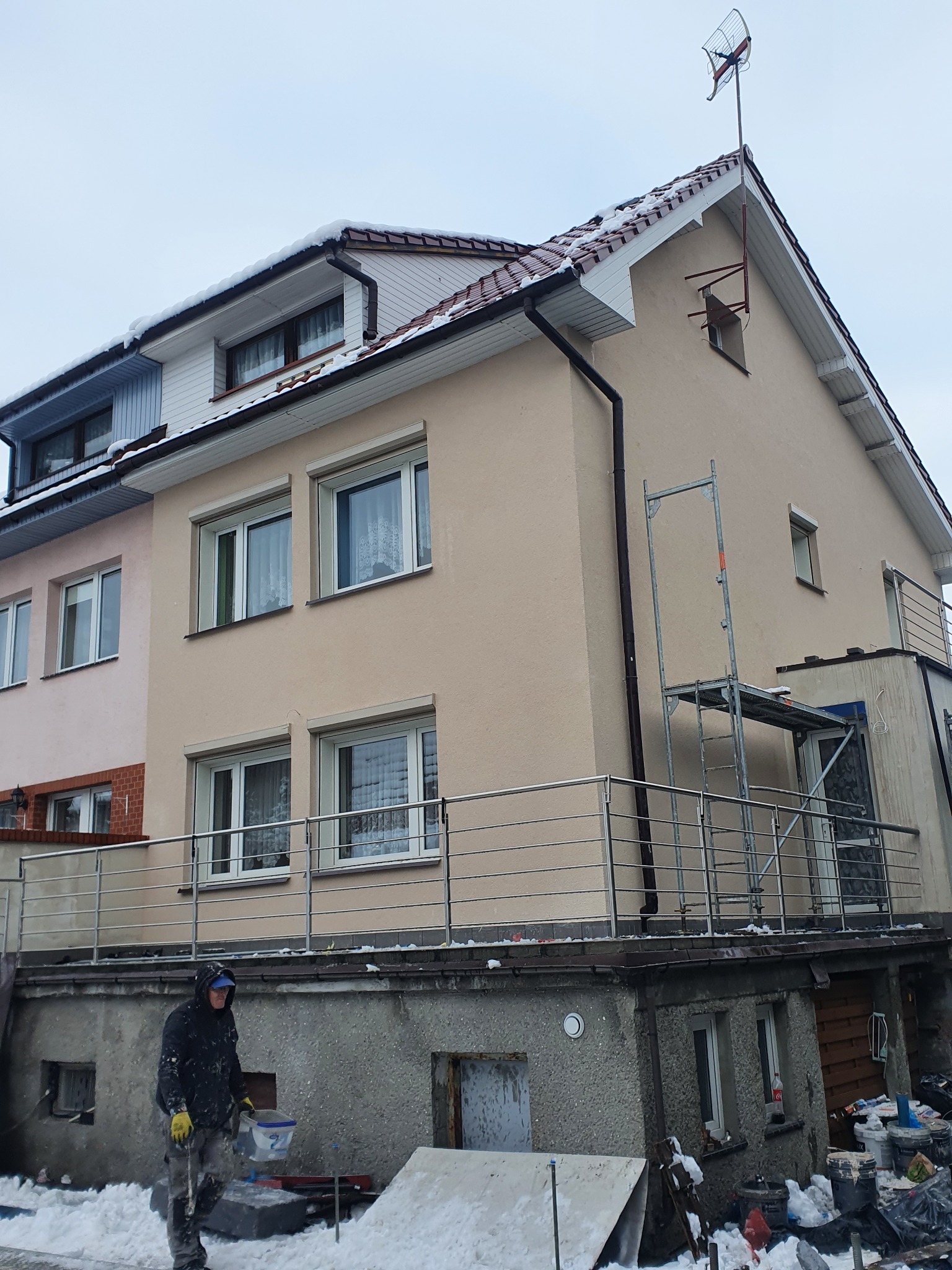Pracownik w zimowej odzieży z wiadrem na tle otynkowanej, beżowej elewacji domu z balkonem i rusztowaniem, widoczny śnieg na ziemi.