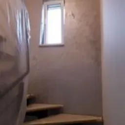 Klatka schodowa w trakcie remontu: surowe drewniane stopnie zabezpieczone folią, ściana z dekoracyjną szarą gładzią, małe okno w białej ramie.