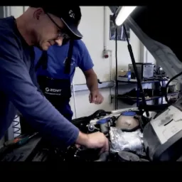 Mechanik w niebieskim kombinezonie firmy Zenit instaluje system zasilania w samochodzie z otwartą maską, widoczny drugi mechanik w tle.