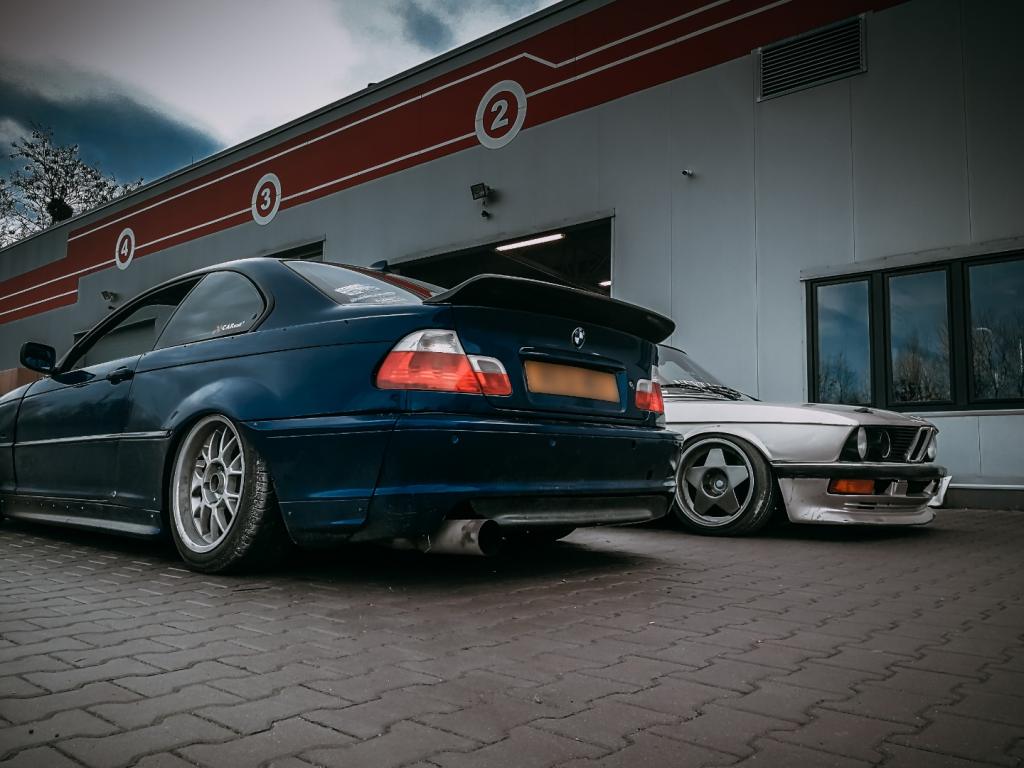 Dwa samochody BMW, ciemnoniebieski E46 z widocznym wydechem i srebrny E28 z felgami w kształcie gwiazdy, zaparkowane przed budynkiem warsztatu z numerami stanowisk.