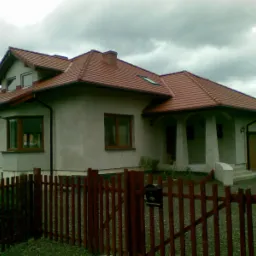 Jednopiętrowy dom z czerwonym dachem, widoczny zza drewnianego ogrodzenia z metalową skrzynką pocztową, pochmurne niebo w tle.