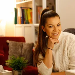 Uśmiechnięta kobieta w kremowym swetrze siedzi w fotelu, opierając brodę na dłoni, w tle regał z segregatorami i czerwony fotel z poduszką.