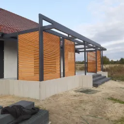 Nowoczesna konstrukcja tarasowa z drewnianymi panelami lamelowymi i szarą metalową ramą, przylegająca do budynku z czerwonym dachem, widoczna w trakcie budowy na tle jesiennego krajobrazu.