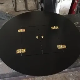 Czarny, okrągły stół z metalu z centralnie umieszczonymi drzwiczkami zamykanymi na złote zawiasy i gałki, widoczne ślady pracy na stole warsztatowym w tle.