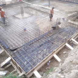 Zbrojenie i szalunek pod płytę fundamentową, widoczni pracownicy na budowie, teren nierówny.