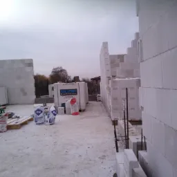 Wznoszenie ścian budynku z bloczków silikatowych, widoczne worki z zaprawą Atlas Silmur i palety z bloczkami Sil-Pro, sznur murarski naprężony wzdłuż ściany, pochmurne niebo w tle.