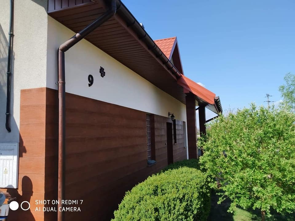 Elewacja domu jednorodzinnego z drewnopodobną okładziną, brązowe rynny i czerwony dach, numer domu '95' na białej ścianie, widok z ogrodu w słoneczny dzień.