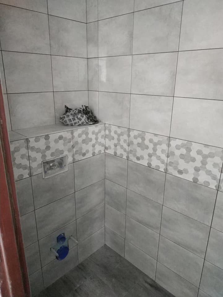 Wykończona kabina prysznicowa z szarymi płytkami, dekoracyjnym pasem z motywem kwiatowym i ukrytą spłuczką, widoczne niebieskie elementy instalacji wodnej, na półce leży szmatka.