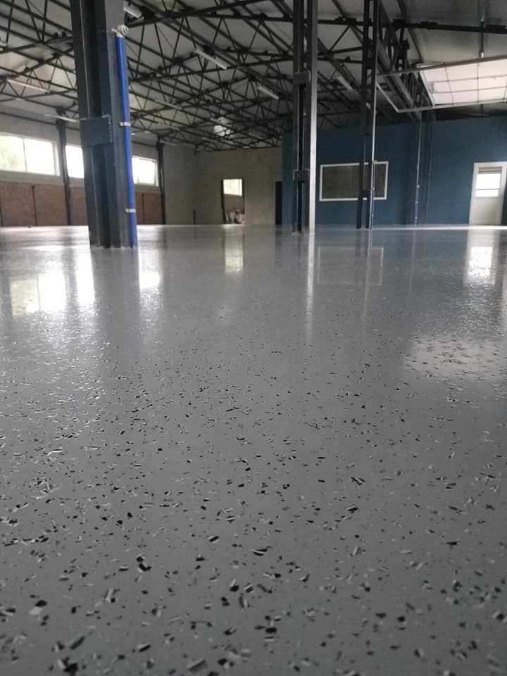 Błyszcząca, szara posadzka epoksydowa z czarnymi drobinkami w pustym, przemysłowym wnętrzu z metalową konstrukcją dachu i niebieską ścianą w tle.