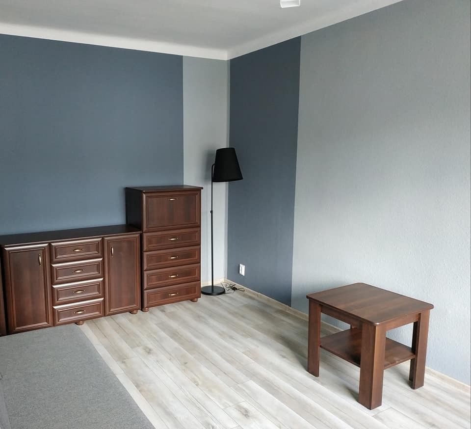 Pokój po remoncie: ściany pomalowane na dwa odcienie szarości i niebieskiego, brązowe meble w stylu retro, drewniana podłoga i czarna lampa stojąca.