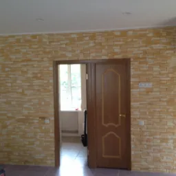 Ściana wykończona panelami imitującymi cegłę w odcieniach beżu i brązu, widoczna obok brązowych drzwi wewnętrznych i białej lodówki.