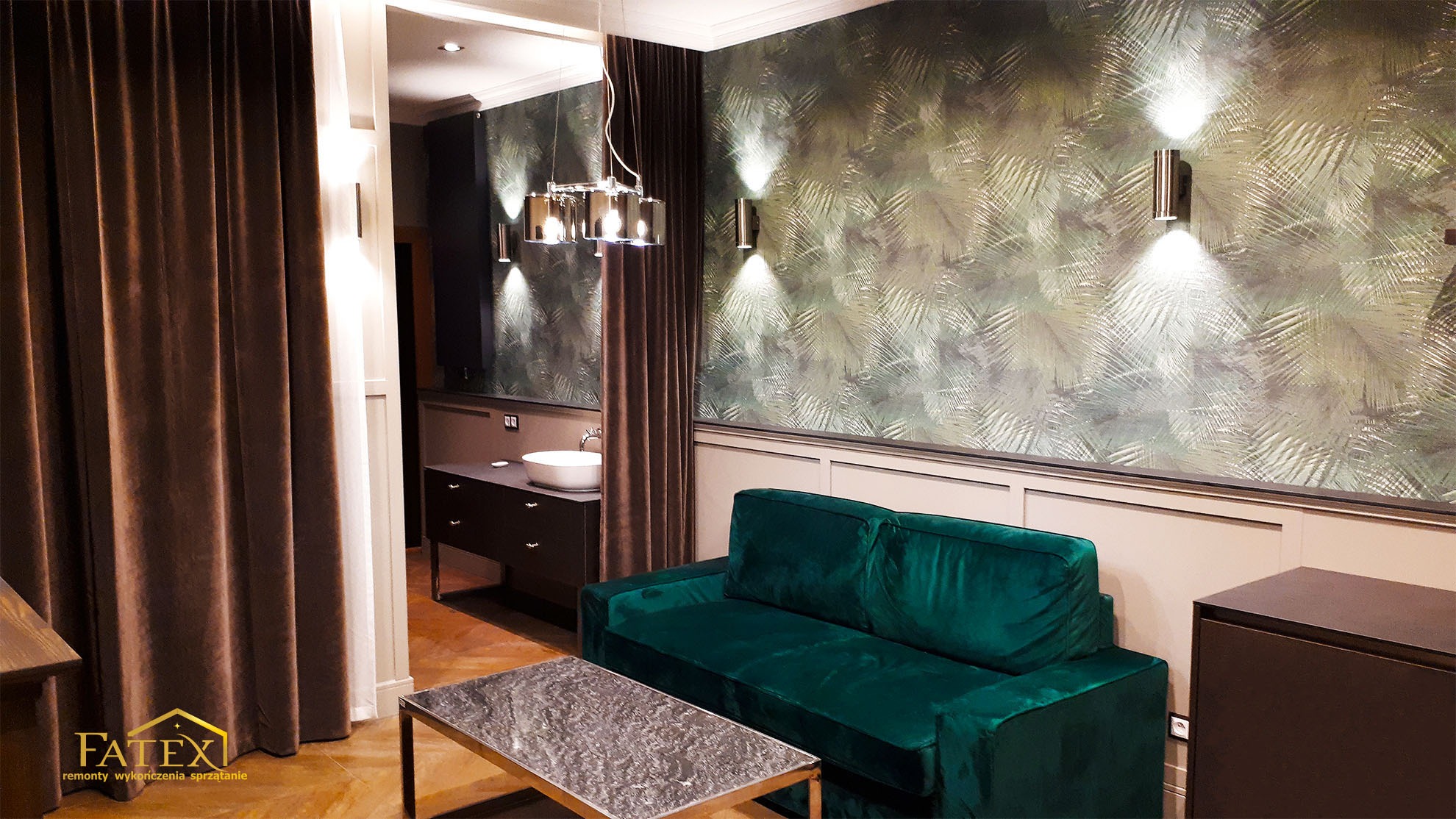 Elegancki salon z zieloną sofą, wzorzystą tapetą w odcieniach zieleni i beżu, oraz nowoczesnym oświetleniem punktowym i wiszącą lampą. W tle widoczna łazienka z umywalką nablatową i ciemnymi meblami.