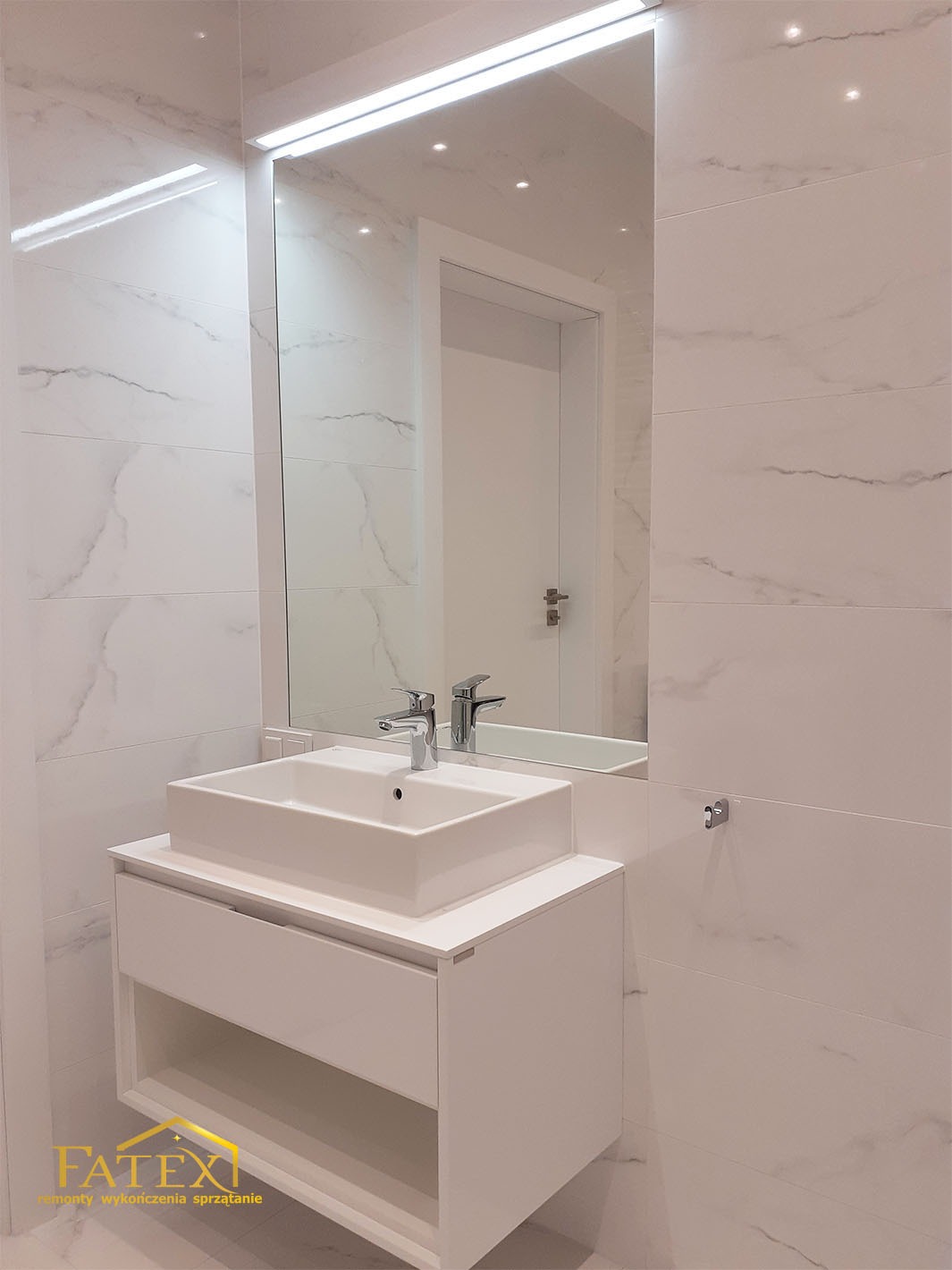 Nowoczesna łazienka z białą umywalką nablatową, szafką pod umywalką z otwartą półką, lustrem z podświetleniem LED i płytkami imitującymi marmur na ścianach.