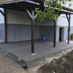 Zadaszony taras z ciemnymi, drewnianymi słupami i szarą podłogą z kostki, widoczny system odwadniający i sprzęt czyszczący w tle.