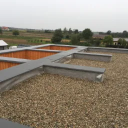 Płaski dach pokryty drobnym żwirem z drewnianą obudową i szarą obróbką blacharską, widok na pola uprawne i zabudowania w tle.