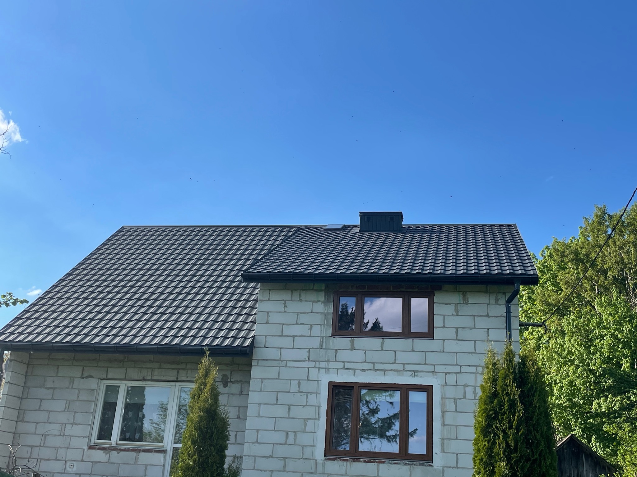 Dom w trakcie budowy z nowym, grafitowym dachem z blachodachówki modułowej, czarnymi rynnami i kominem, okna z brązowymi ramami, ściany z białego betonu komórkowego, iglaki przed budynkiem...