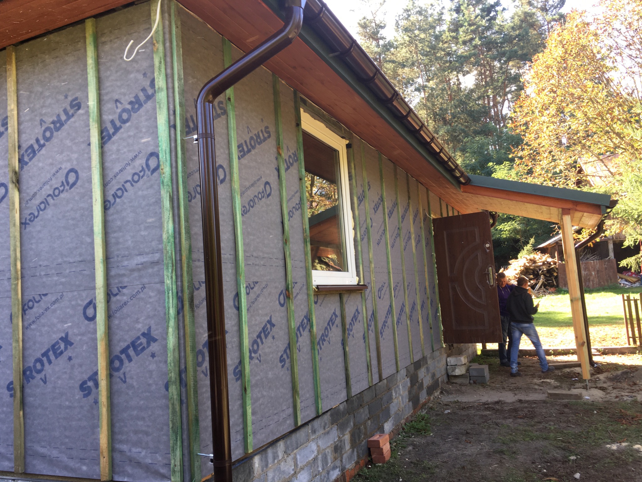 Dom w trakcie ocieplania, widoczne drewniane łaty, membrana wiatroizolacyjna z nadrukiem, zamontowana rynna, okno i częściowo wykończony dach. W tle drzewa i osoby wchodzące do budynku.