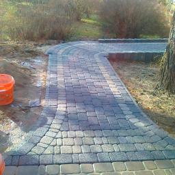 Hydro-Lux Mariusz Śliczniak - Zakrzywiony chodnik z szarej kostki brukowej prowadzący przez ogród, widoczne wiadro i narzędzia, częściowo ukończony fragment.