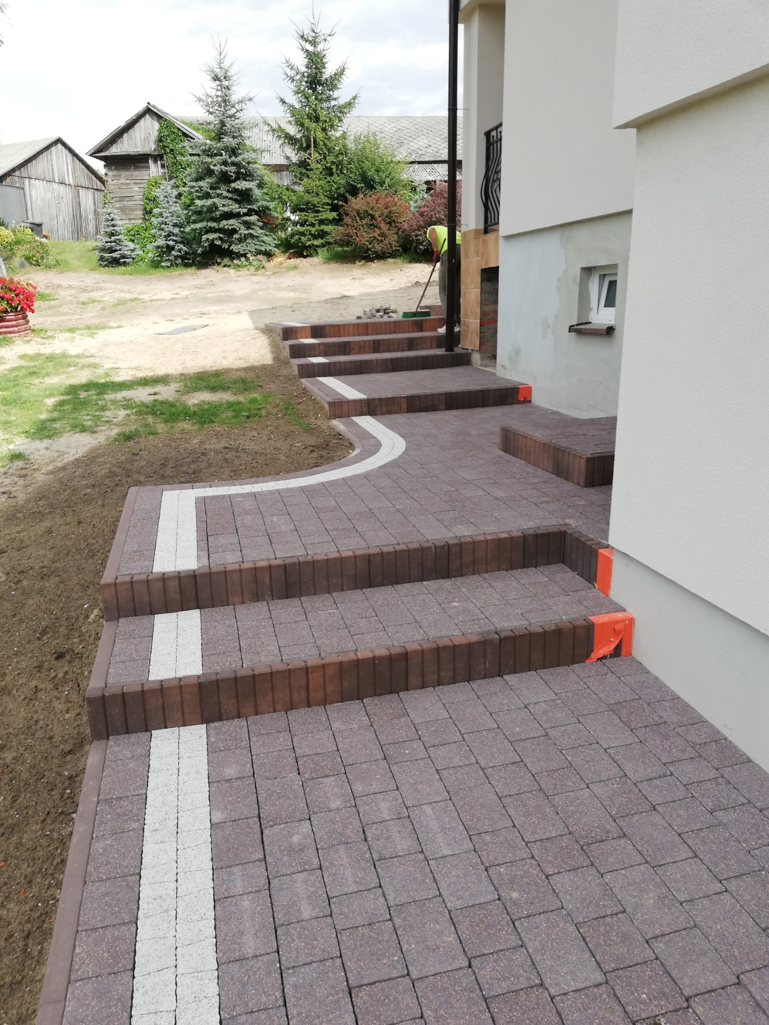 Schody wejściowe do domu wyłożone brązową kostką brukową z białymi akcentami, widoczne zabezpieczenia na krawędziach stopni i pracownik z miotłą w tle.