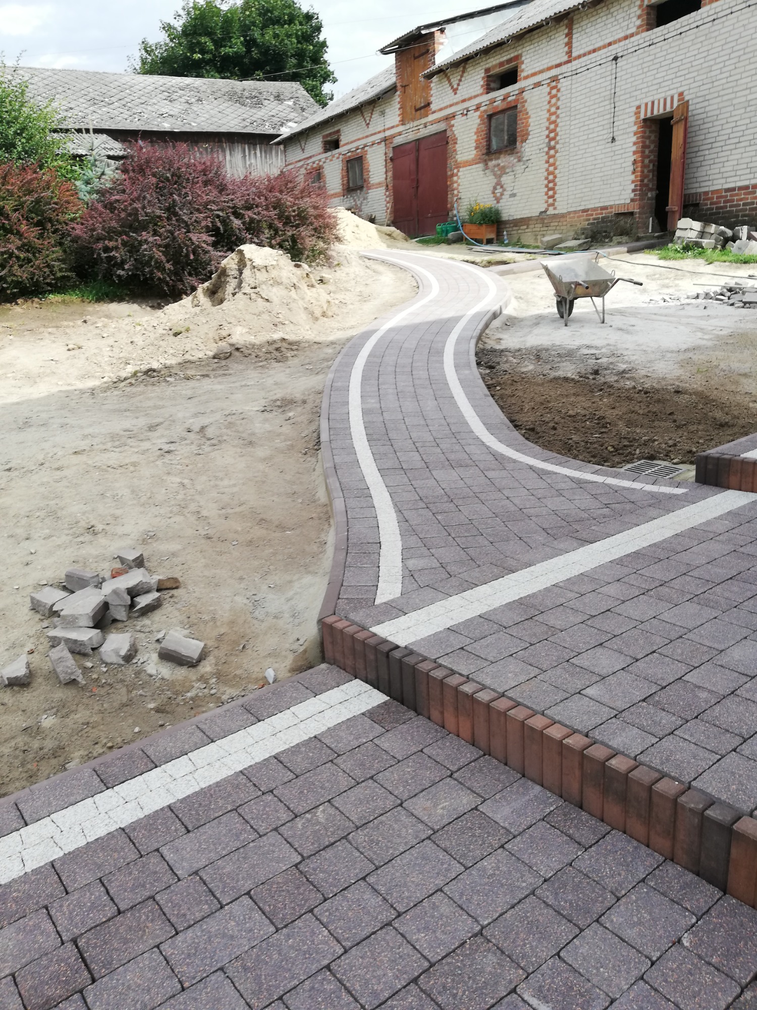 Ścieżka z kostki brukowej w kształcie litery S, obrzeża z czerwonej kostki, białe pasy oddzielające, w tle budynki gospodarcze i kupa piachu, widok z góry.