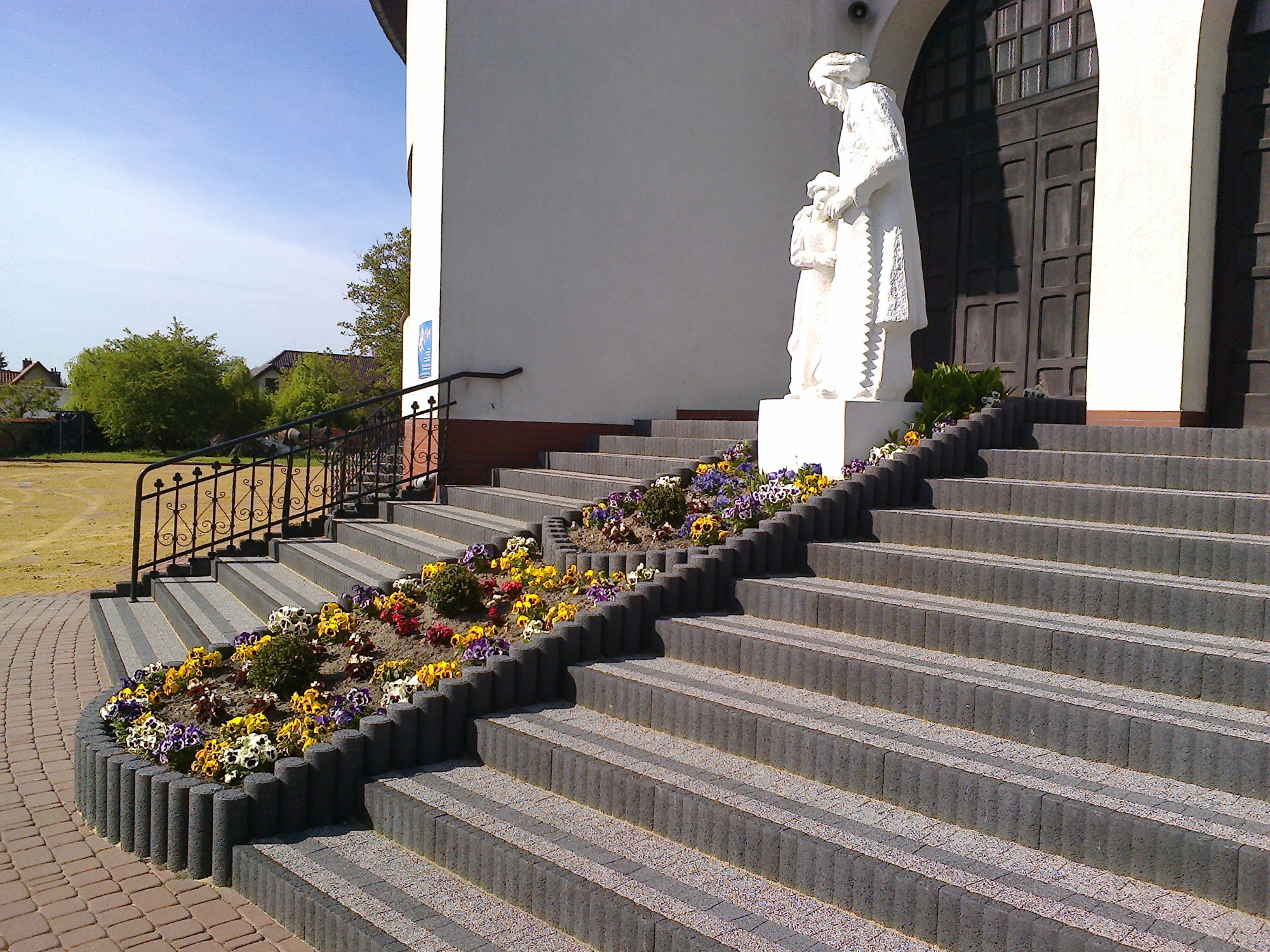 Schody wejściowe do budynku w Wólce wykończone kostką brukową z obrzeżami rabat kwiatowych, prowadzące do posągu.
