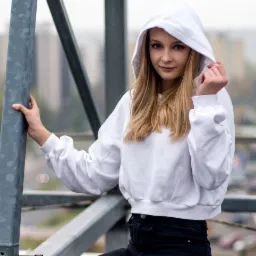 Portret młodej kobiety w białej bluzie z kapturem, opierającej się o metalową konstrukcję na tle miejskiego krajobrazu.