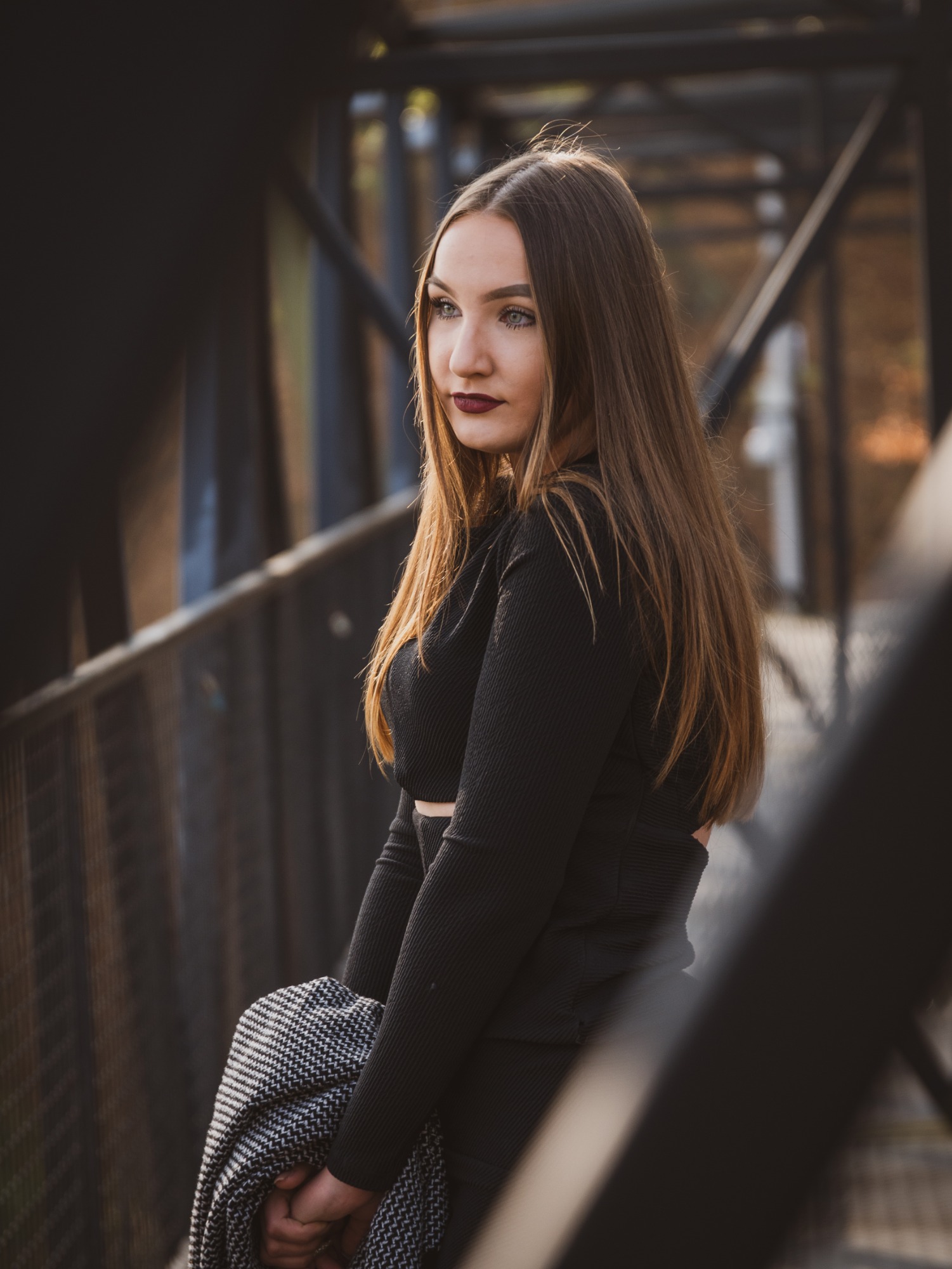 Portret kobiety o długich brązowych włosach, w czarnym ubraniu, stojącej na metalowym moście i spoglądającej w bok, trzymającej w rękach wzorzysty szal.