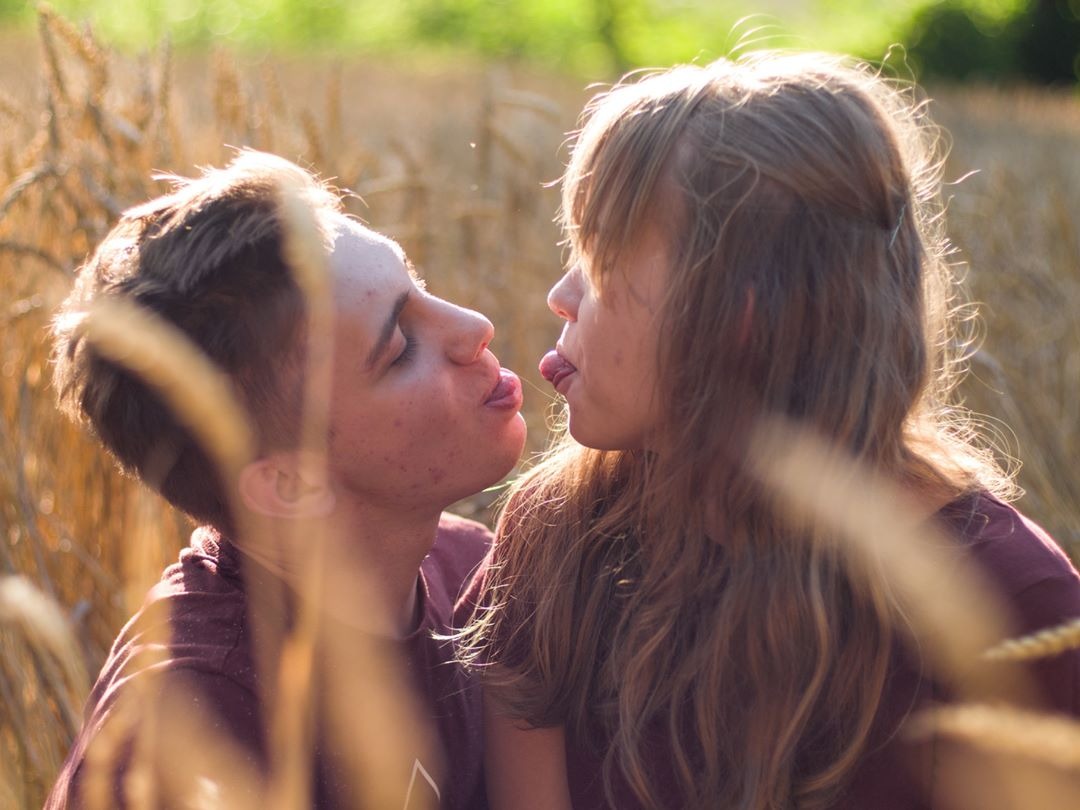 Młoda para w polu pszenicy, wyciągnięte języki, ciepłe światło zachodzącego słońca, delikatny bokeh w tle.