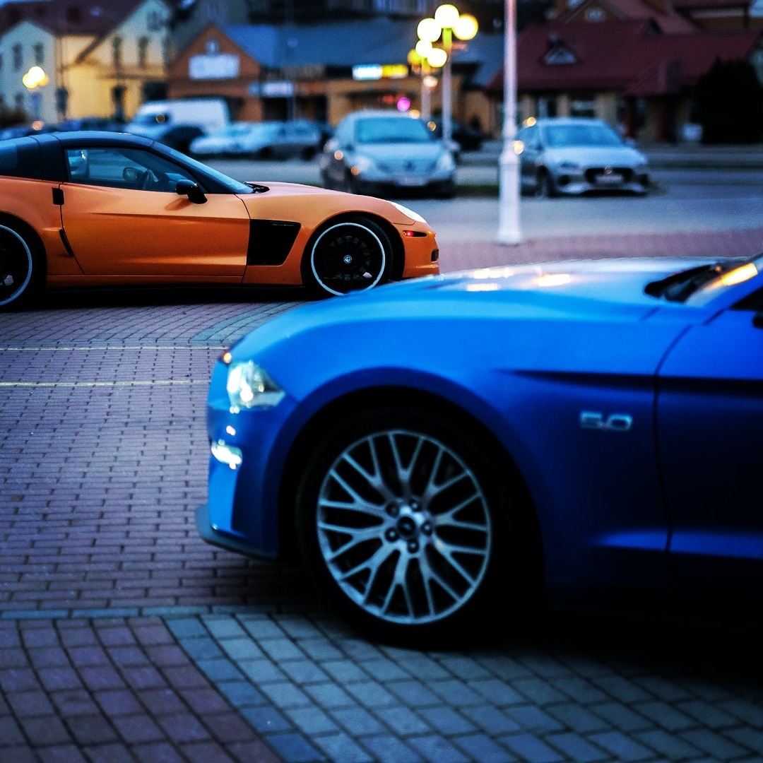 Pomarańczowy Chevrolet Corvette obok niebieskiego Forda Mustanga na brukowanej ulicy, z innymi samochodami i budynkami w tle o zmierzchu.