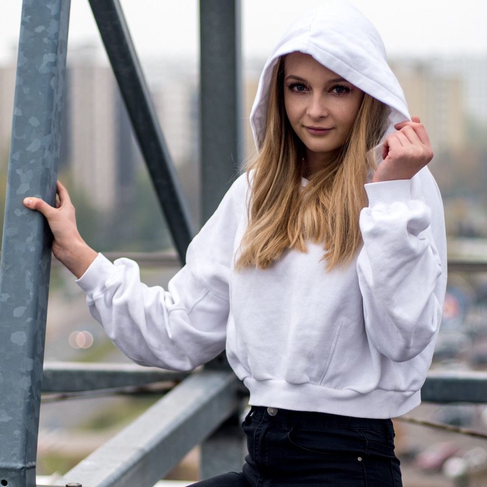 Portret młodej kobiety w białej bluzie z kapturem, opierającej się o metalową konstrukcję na tle miejskiego krajobrazu.