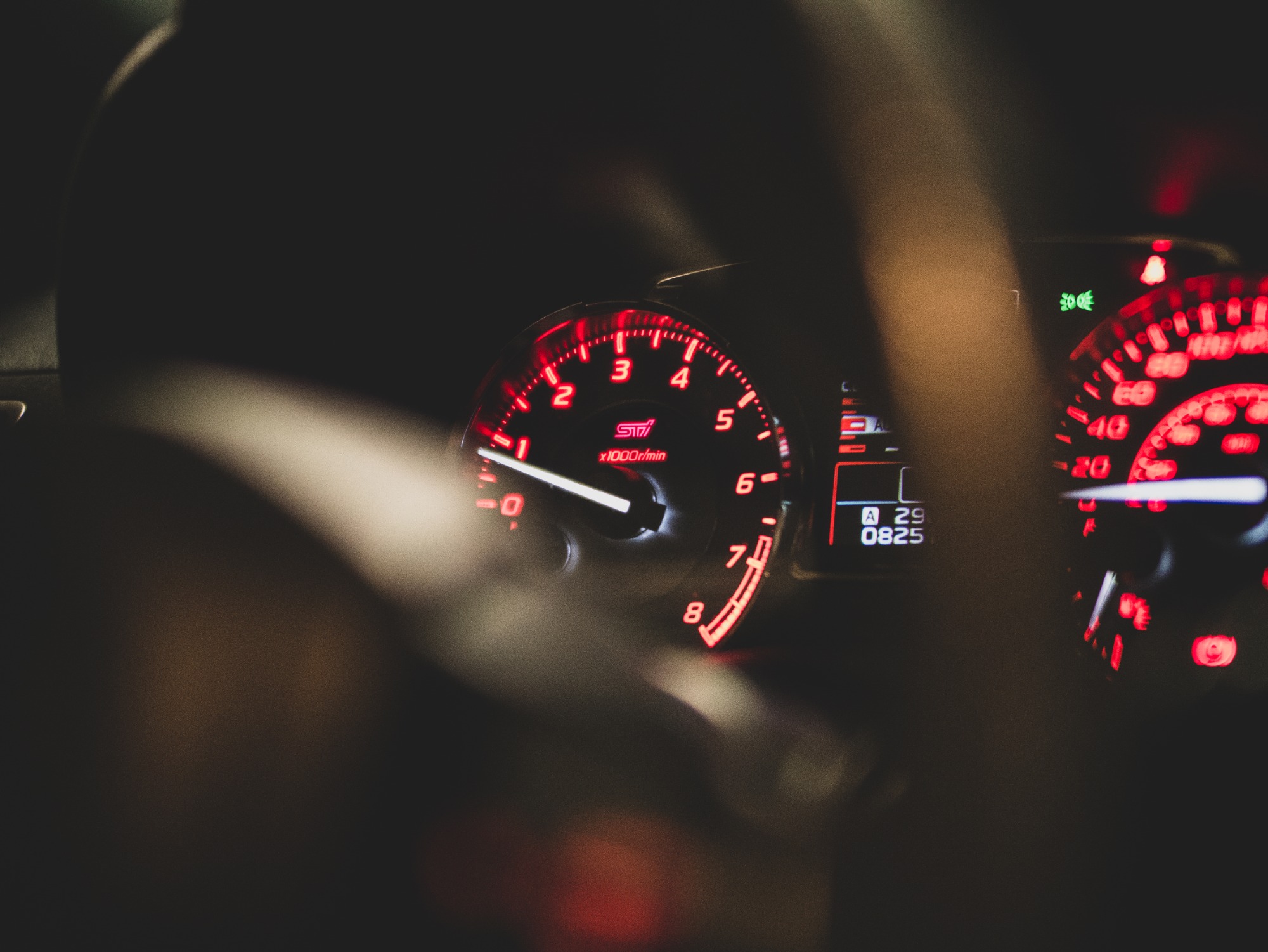 Podświetlony na czerwono licznik obrotów w samochodzie marki Subaru STI, wskazujący około 1500 obrotów na minutę, widoczny przez kierownicę w ciemnym wnętrzu pojazdu.