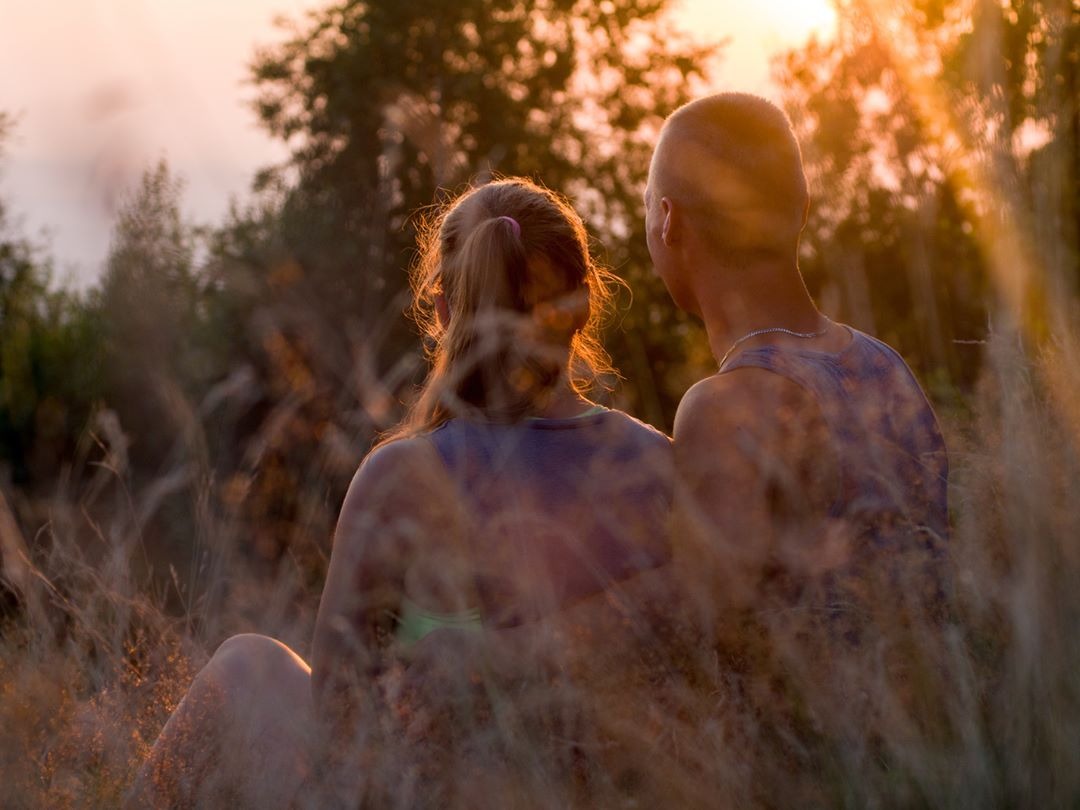 Para siedzi w trawie, odwrócona tyłem, oświetlona zachodzącym słońcem, widok przez źdźbła trawy.