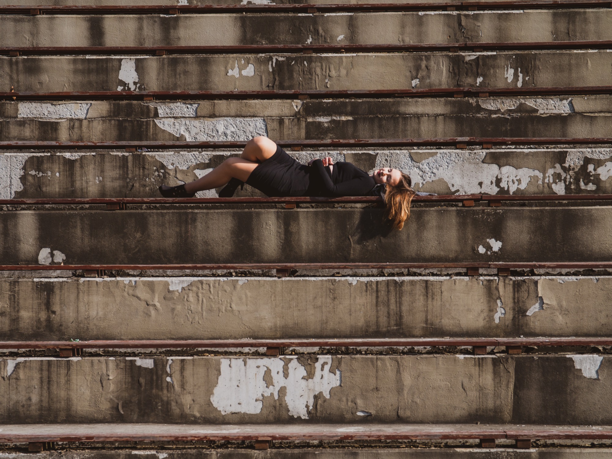 Kobieta w czarnej sukience i butach na obcasie leży na betonowych schodach z odpadającą farbą, patrząc w górę.