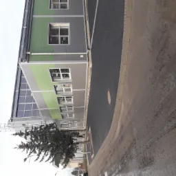 Nowoczesny budynek przemysłowy z szaro-zielonymi płytami warstwowymi, czarną kostką brukową i młodym drzewkiem iglastym, sfotografowany w pochmurny dzień z perspektywy chodnika.