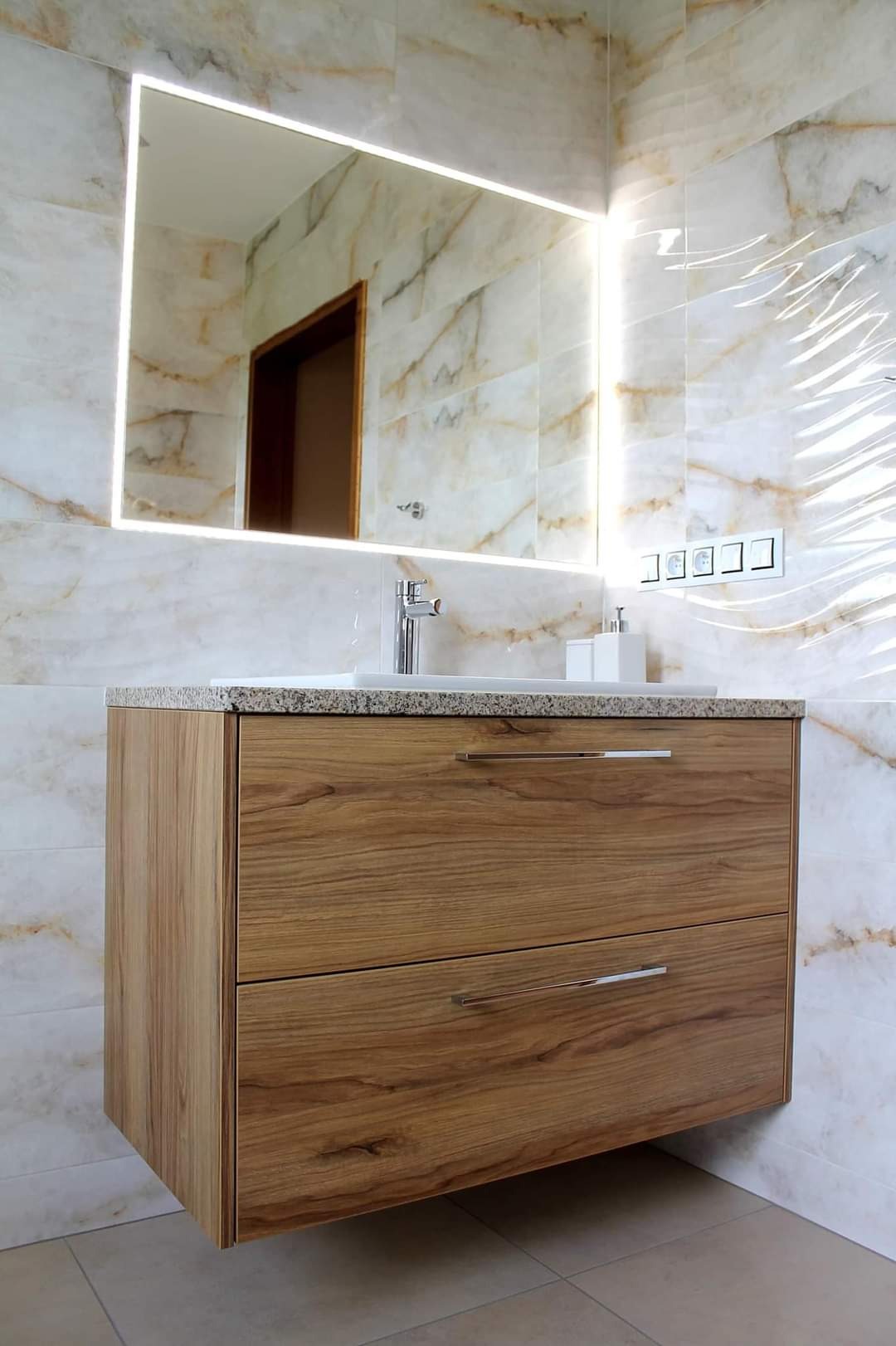 Nowoczesna łazienka z podwieszaną szafką z drewnianym fornirem, granitowym blatem i lustrem z podświetleniem LED, płytki marmurowe na ścianach.