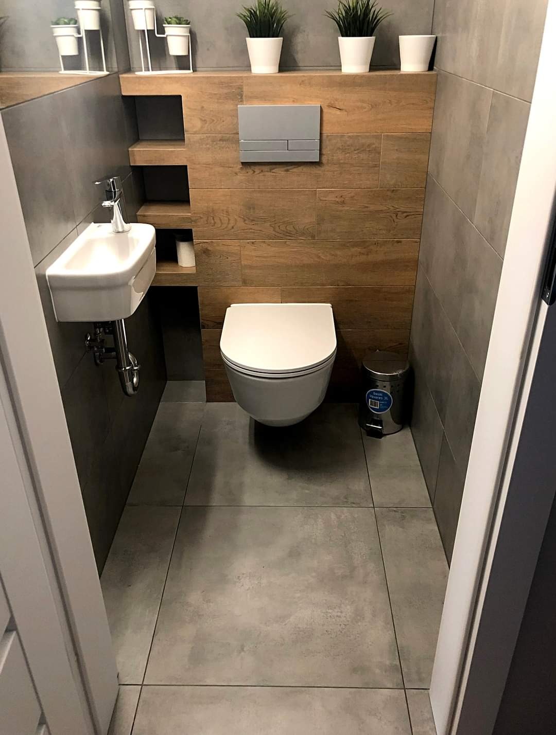 Nowoczesna, wąska łazienka z szarymi płytkami podłogowymi i ściennymi, drewnopodobną ścianą z zabudowaną spłuczką, wiszącą umywalką i toaletą oraz metalowym koszem na śmieci.