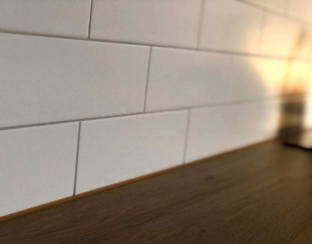 Białe, prostokątne płytki ułożone na ścianie nad blatem kuchennym o drewnianej fakturze, widoczne delikatne spoiny i ciepłe światło w tle.