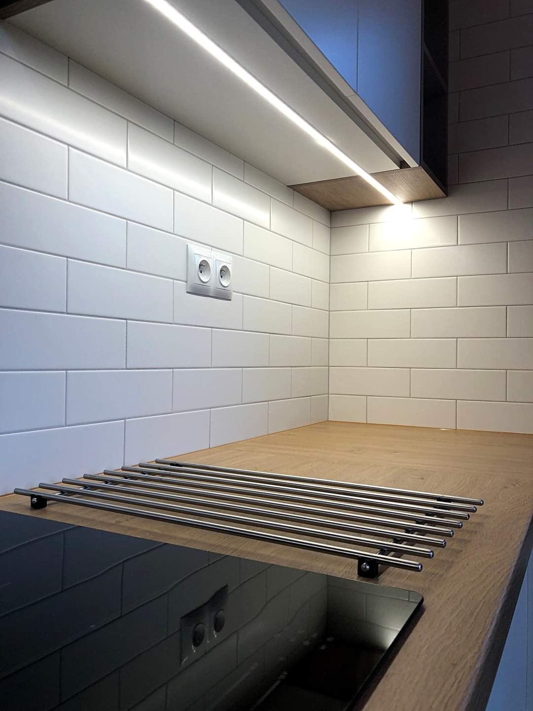 Białe, prostokątne płytki na ścianie nad drewnianym blatem kuchennym, oświetlenie LED podszafkowe i metalowy ociekacz nad czarną płytą indukcyjną.