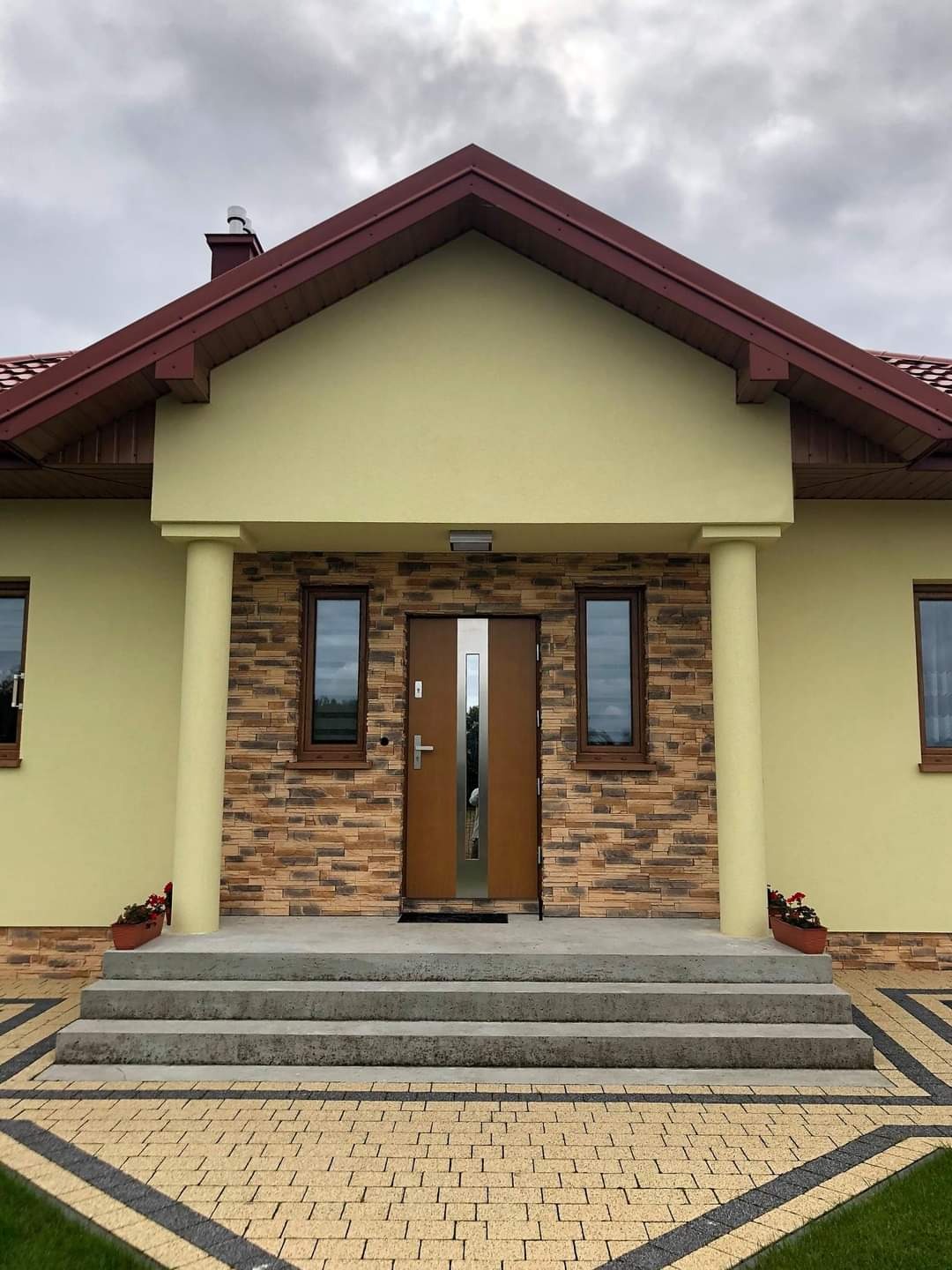 Elewacja domu z okładziną z kamienia dekoracyjnego na ścianie frontowej, brązowe okna i drzwi wejściowe, schody wejściowe z kostki brukowej w kolorze żółtym i czarnym