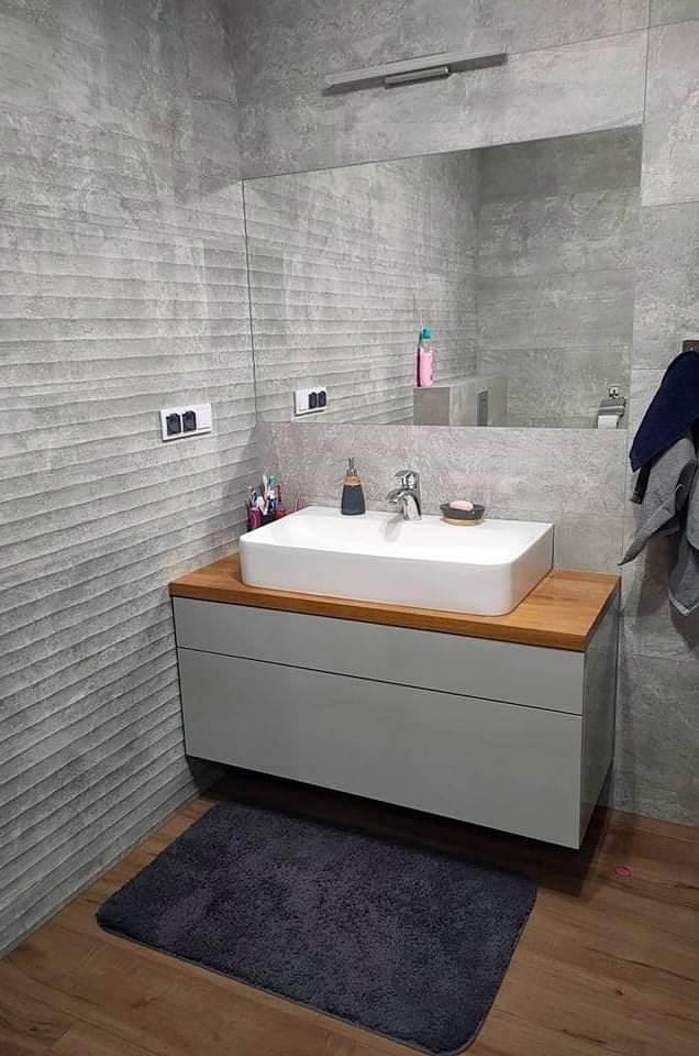 Nowoczesna łazienka z szarymi płytkami imitującymi beton, białą umywalką nablatową na drewnianym blacie i szarą szafką pod umywalką oraz ciemnoszarym dywanikiem.